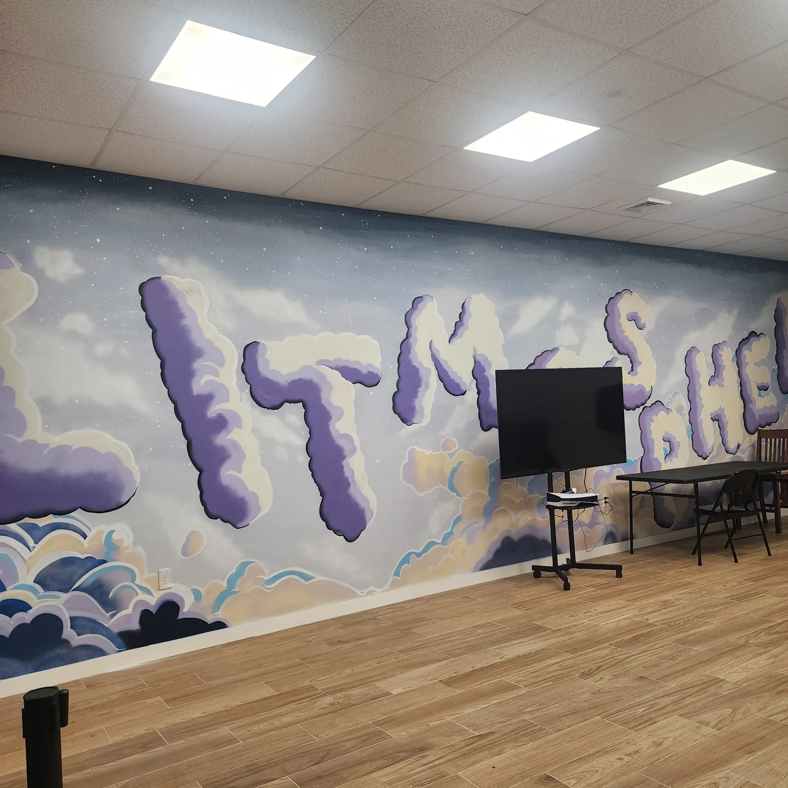 Hand-painted mural of lavender and cream clouds that spell out Litmosphere across the length of the wall, with a screen on a rolling stand and a folding table in front of it
