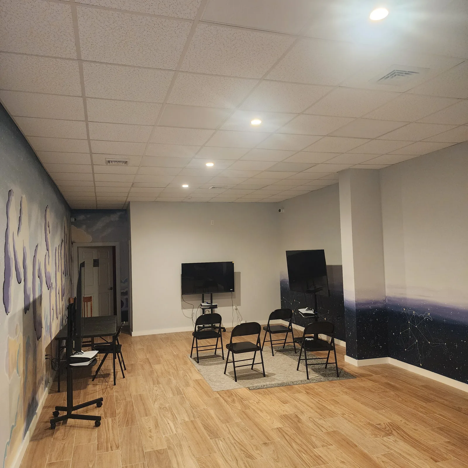 The event space under its recessed ceiling lights, with three screens on stands, a table and folding chairs on a rug